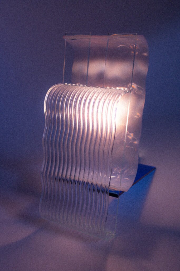 Een witte tafel met een rechtopstaande glazen lamp. De lamp is gemaakt tijdens een glas workshop in utrecht genaamd ''Lamp Maken''. Het glas heeft verschillende kleuren en structuren.