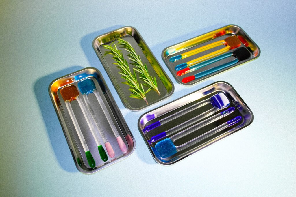 Op een witte tafel liggen verschillende rechthoekige rvs bordjes met hierop glazen roerstaven genaamd ‘pool sticks’. De roerstaven zijn gemaakt van gekleurd glas.