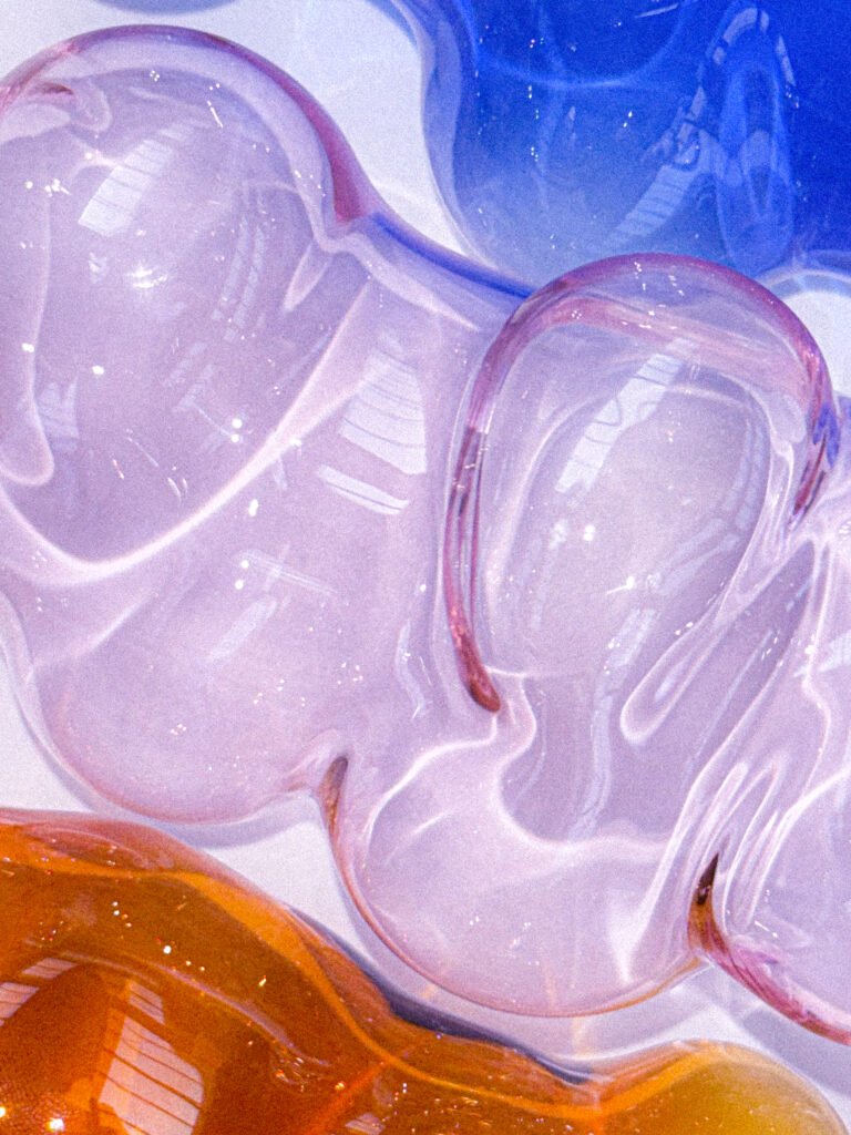 Op de voorgrond liggen drie glazen ornamenten gemaakt van borosilicaatglas. In de kleuren blauw , amber en roze. De vormen zijn organisch en mondgeblazen.